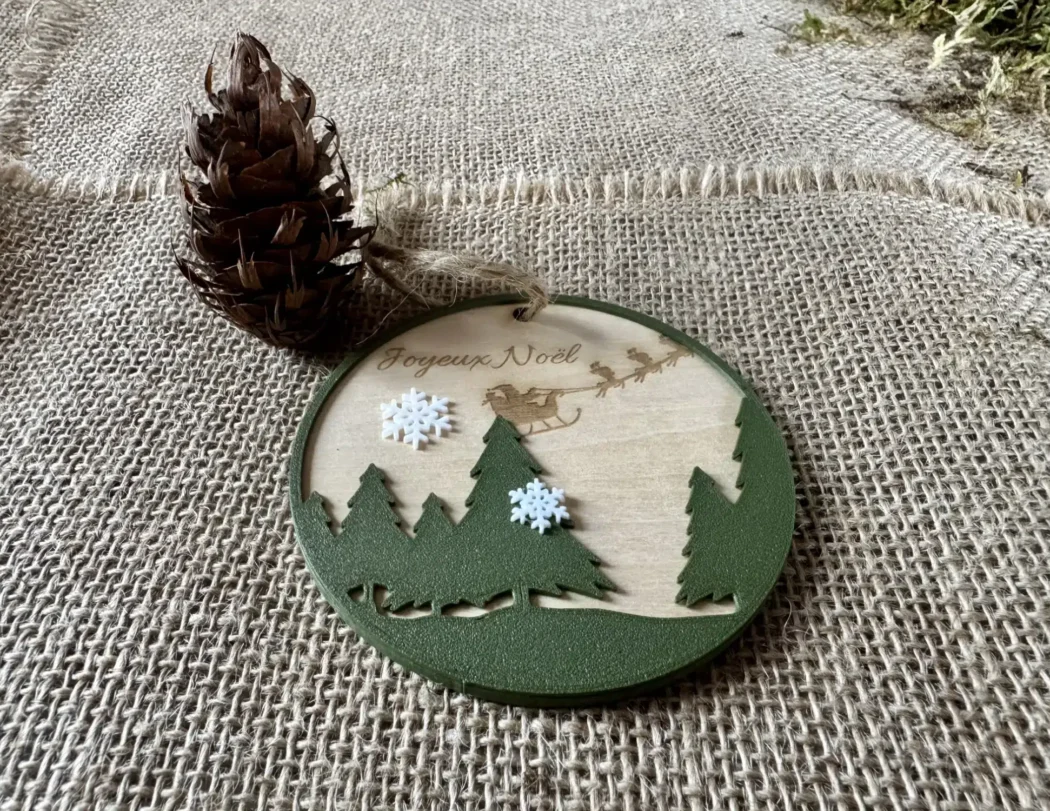 Wooden Christmas ball engraved with the text Merry Christmas, fir trees in the foreground and snowflakes printed in Biodegradable PLA